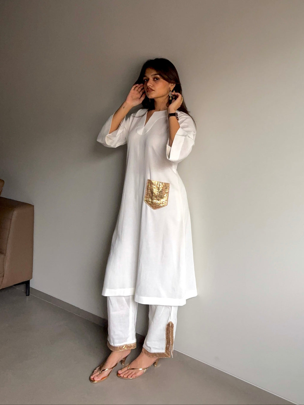 A woman modeling a white kurta set with a metallic gold pocket detail, standing in a minimalist room with a gray wall.