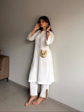 A woman modeling a white kurta set with a metallic gold pocket detail, standing in a minimalist room with a gray wall.